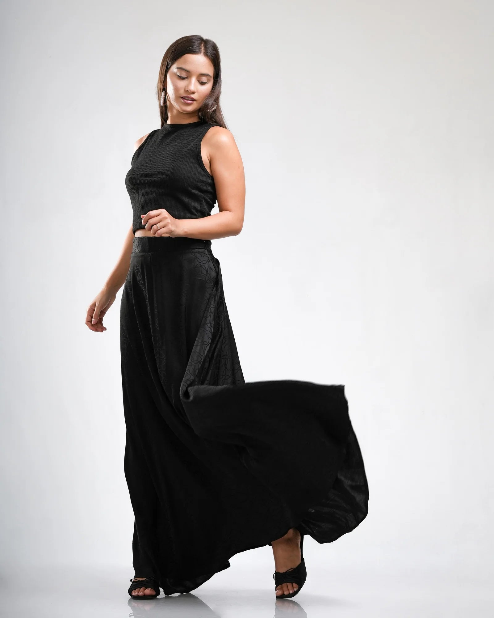 Model in a black sleeveless crop top and a black jacquard-print flowing maxi skirt, caught mid-movement against a pale studio backdrop