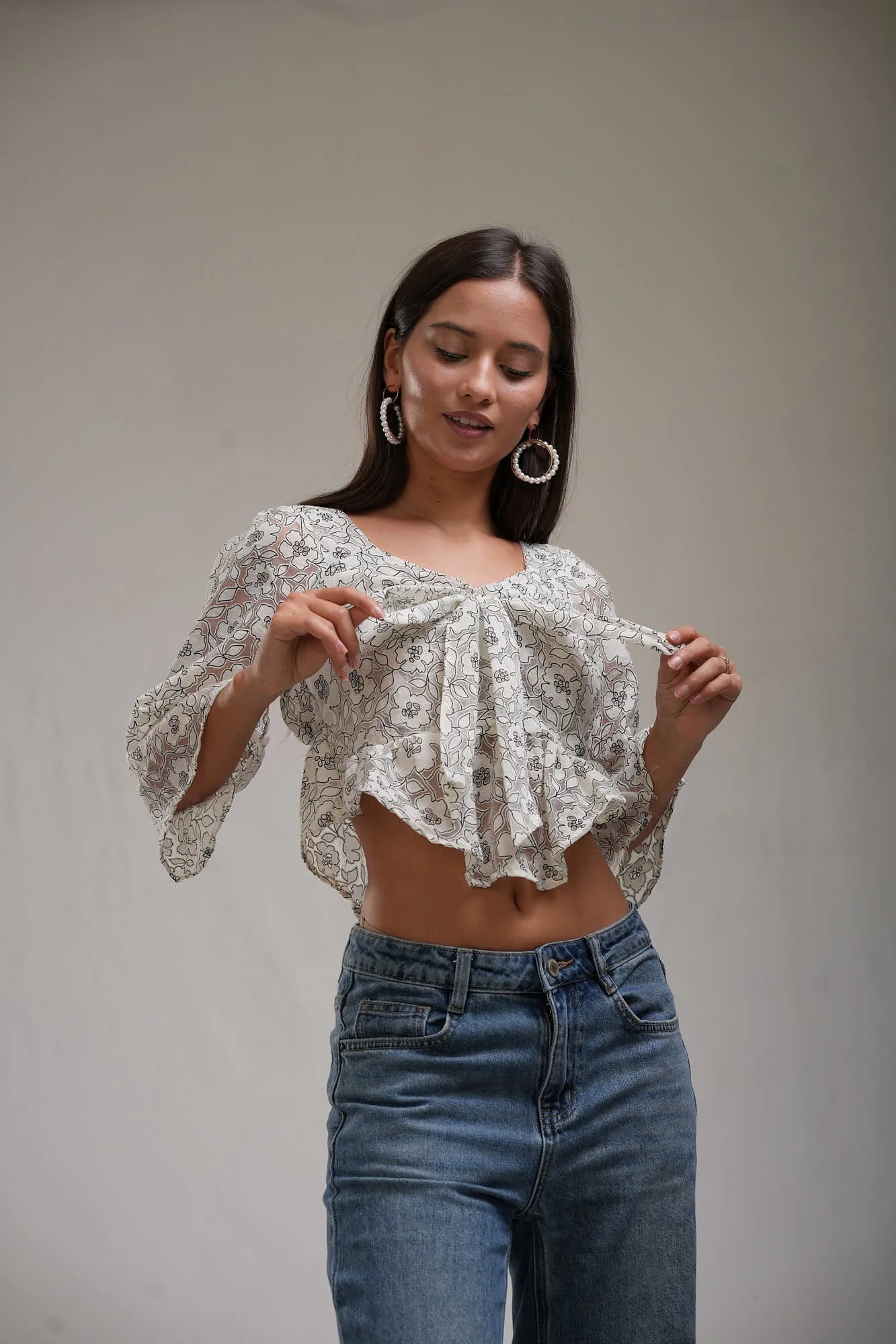 Ivory floral-print crop top with flared sleeves and a ruffled hem, worn with blue wide-leg jeans against a pale studio backdrop