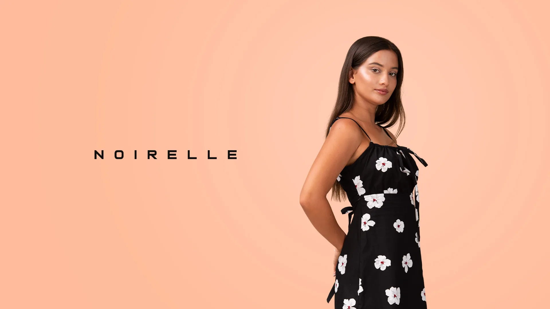 Model in a black floral cami-strap midi dress, Noirelle wordmark beside her on a peach backdrop