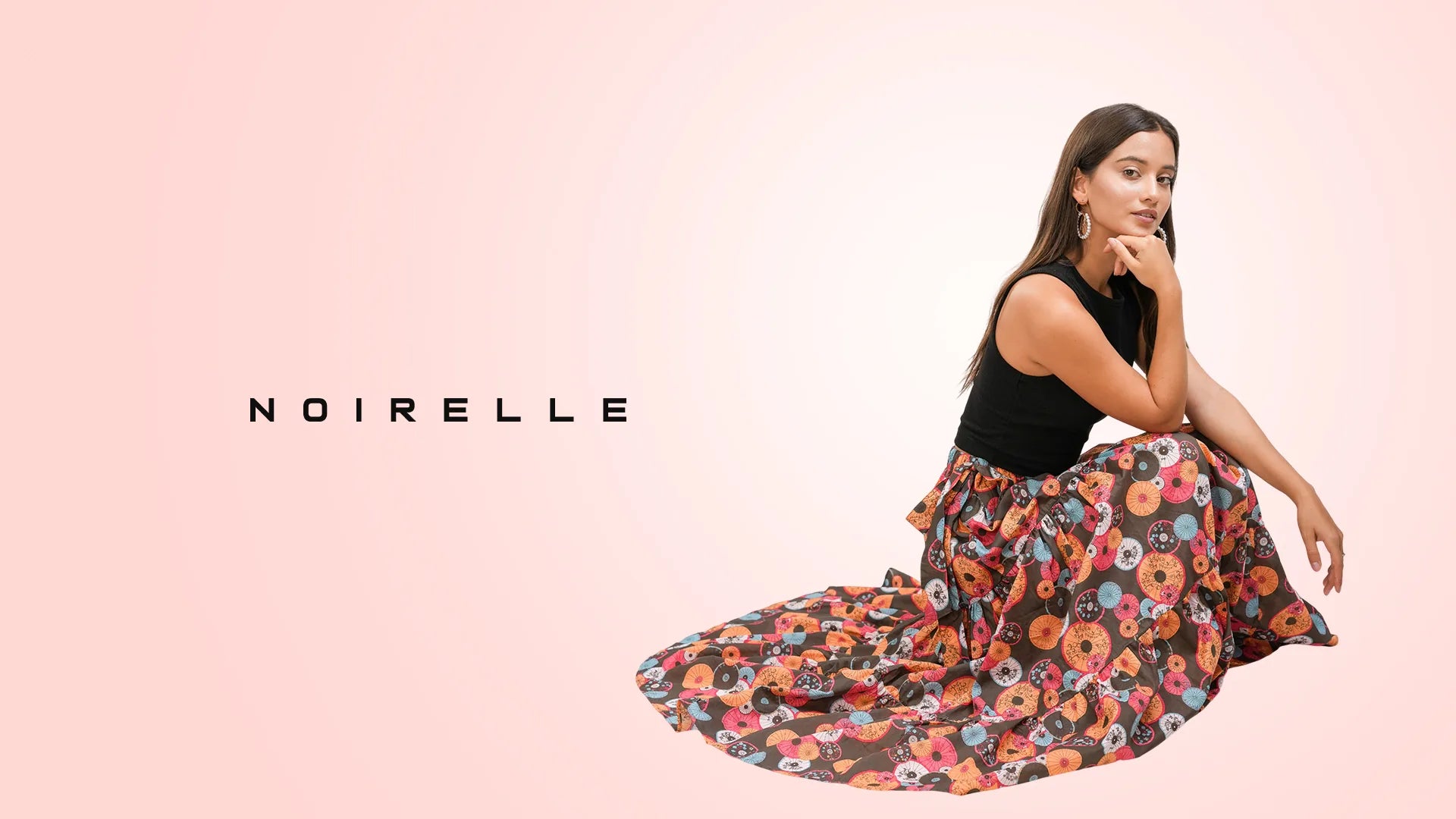 Model seated in a black top and a printed maxi skirt, Noirelle wordmark beside her on a blush backdrop