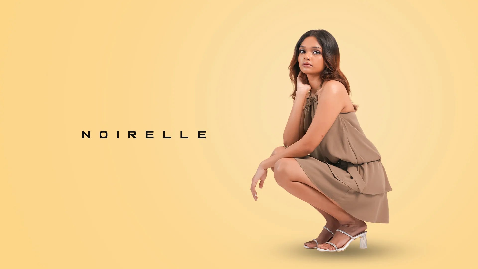 Model crouching in a taupe tiered mini dress, Noirelle wordmark beside her on a butter-yellow backdrop