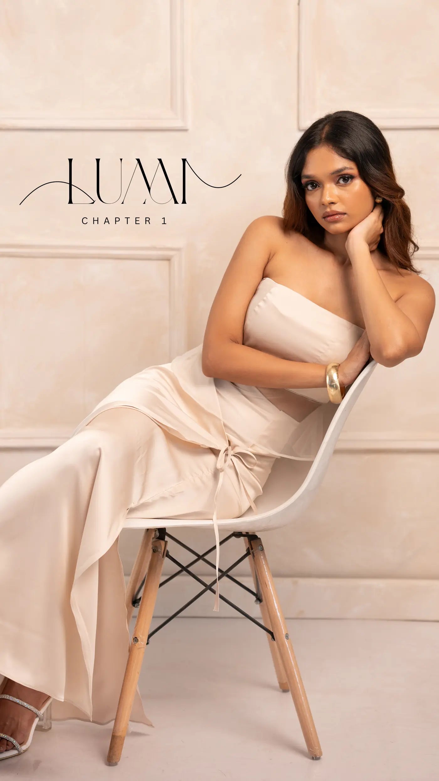 Model seated on a white chair in a champagne satin strapless maxi dress with a tie waist, LUMA Chapter 1 campaign lockup on a cream panelled wall