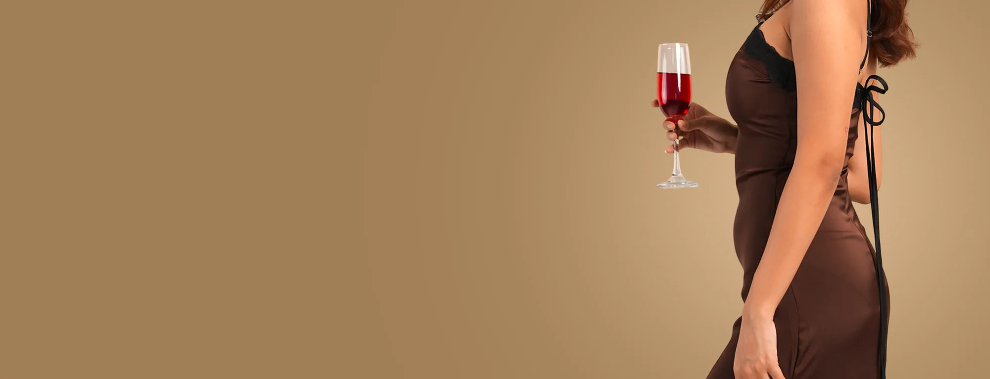 Model in a chocolate-brown satin slip dress with black lace trim and tie back, holding a glass of red wine against a tan backdrop