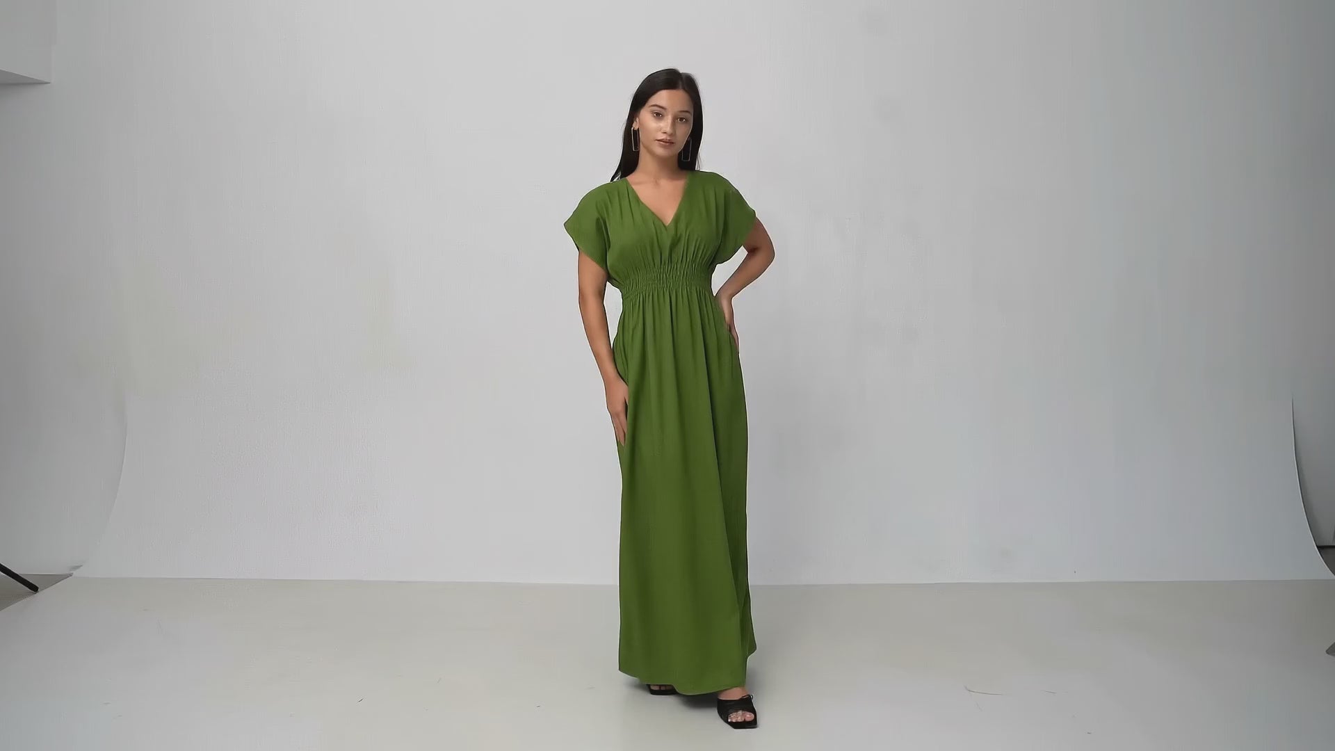 Model turning in an olive-green kaftan-style midi dress with cap sleeves and a smocked waist, against a pale studio backdrop